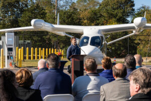 Geoff Douglass tells the story of his instigating the development of electric aircraft charging stations at Marshfield Municipal Airport. BETA Technologies’ developmental electric aircraft ALIA and Level 3 charging station loom behind. Photo courtesy of BETA Technologies